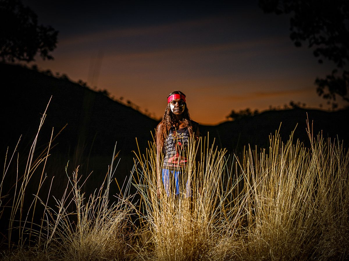 A young woman in traditional face paint stands in grass at sunset.